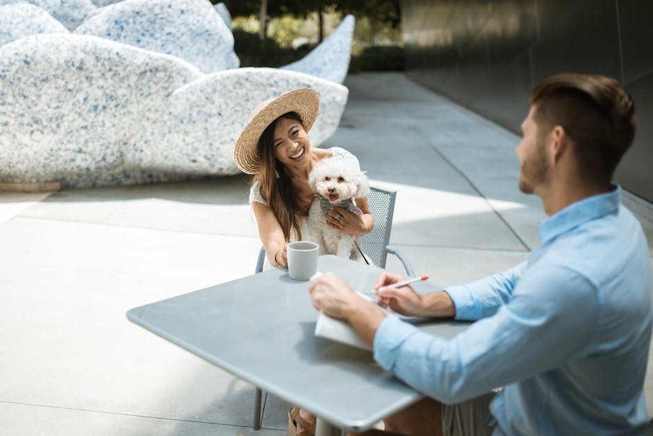 A woman with a straw hat holds her dog at a sunny outdoor cafe table, engaged in a friendly meeting.