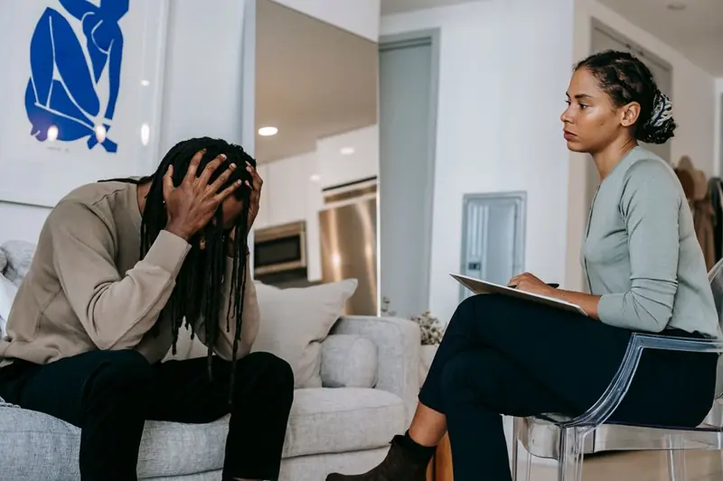 Concentrated African American female psychotherapist in formal wear sitting on chair and consulting desperate male touching head in frustration